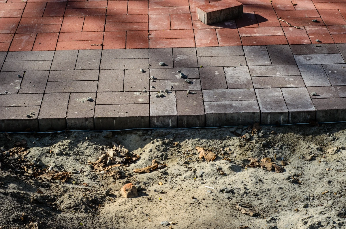 Rozpoczęte układanie kostki brukowej na chodniku, widoczna warstwa piasku i obrzeże z krawężników betonowych