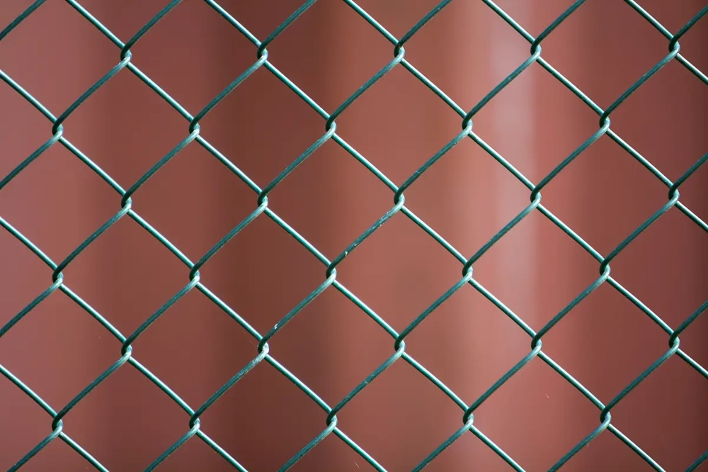 Metalowa siatka ogrodzeniowa z romboidalnym splotem na tle brązowej ściany