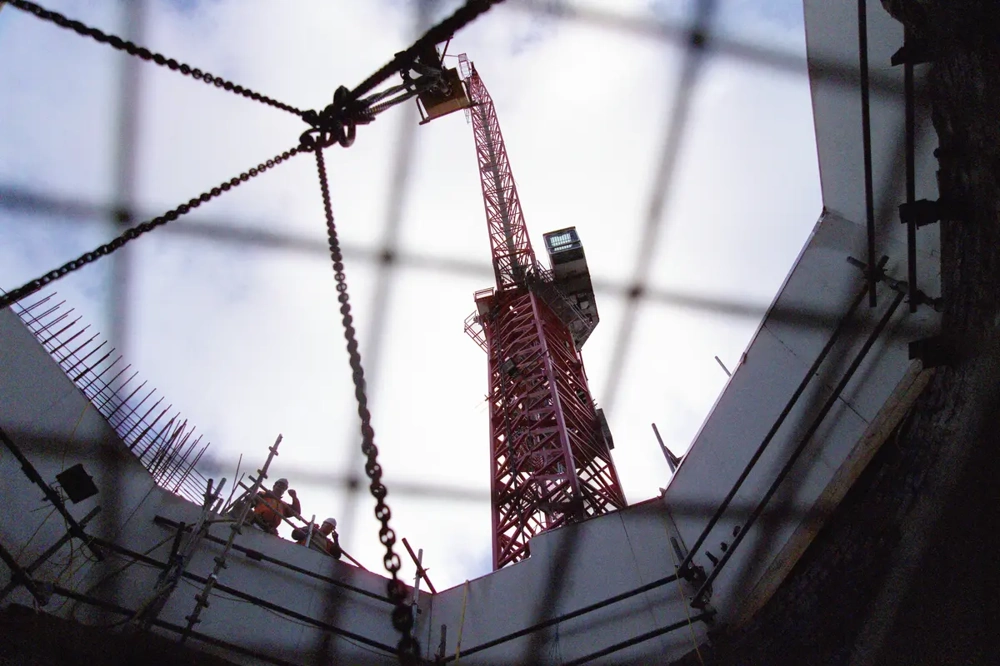 Prace ziemne przy głębokim wykopie z widocznym żurawiem wieżowym i robotnikami